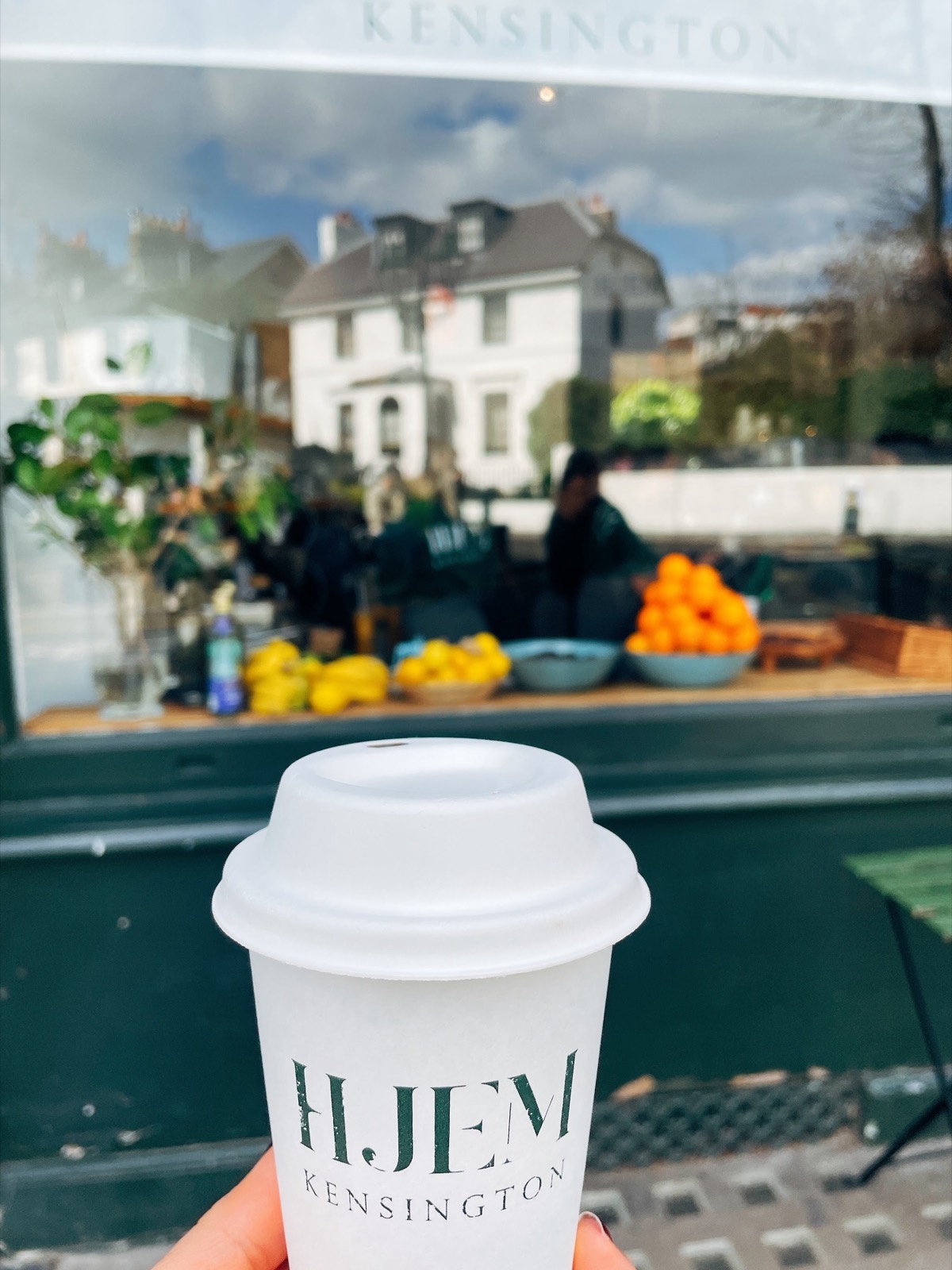 A HJEM takeaway cup held up in front of the dark green Launceston Place shopfront